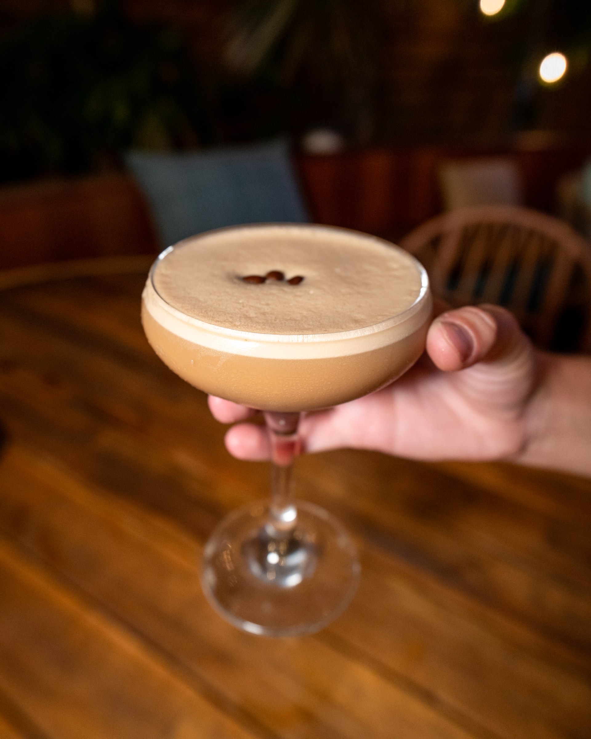 A hand presenting an espresso martini over a wooden restaurant table