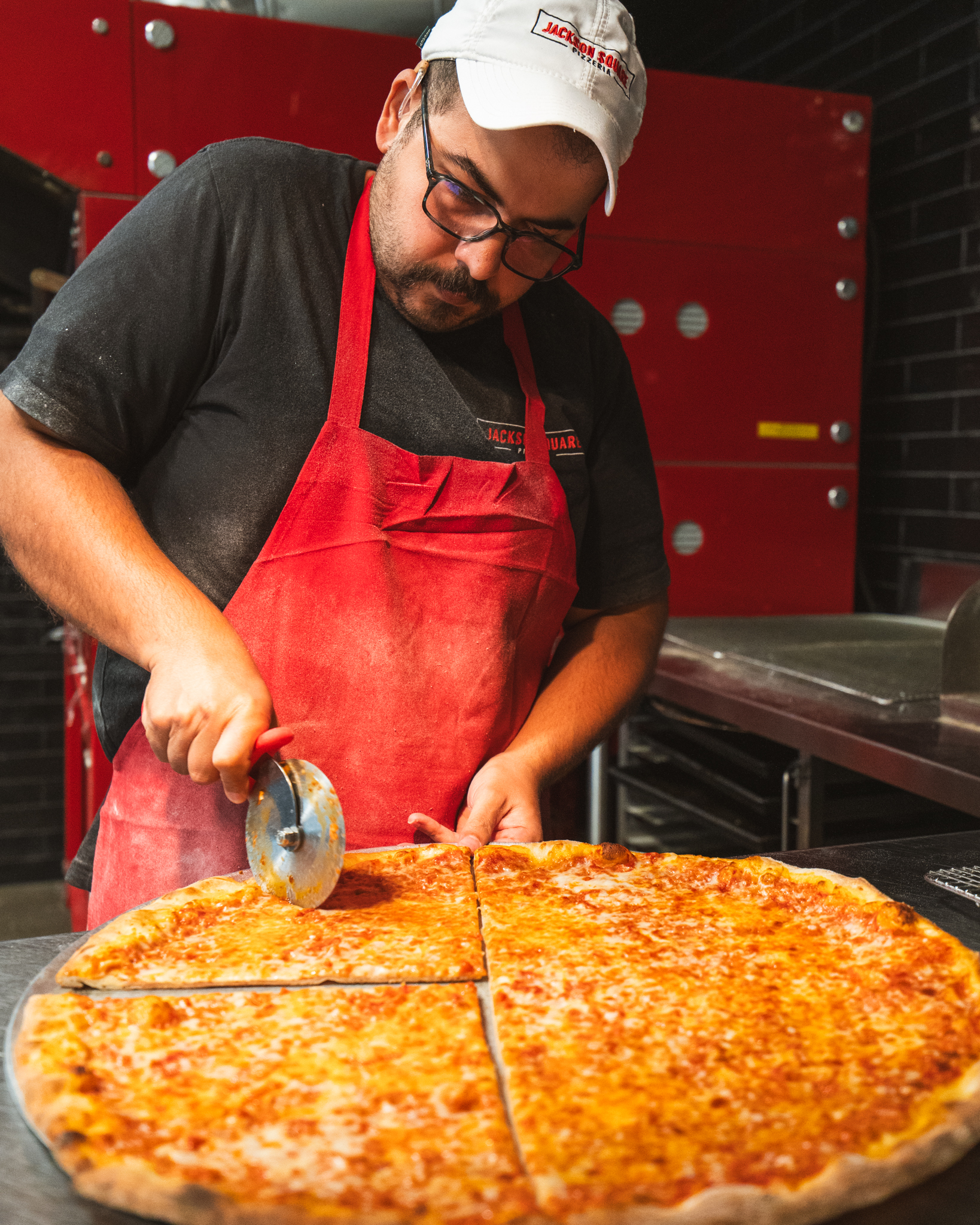 A Jackson Square pizza maker in a red apron cutting a cheese pizza at the counter