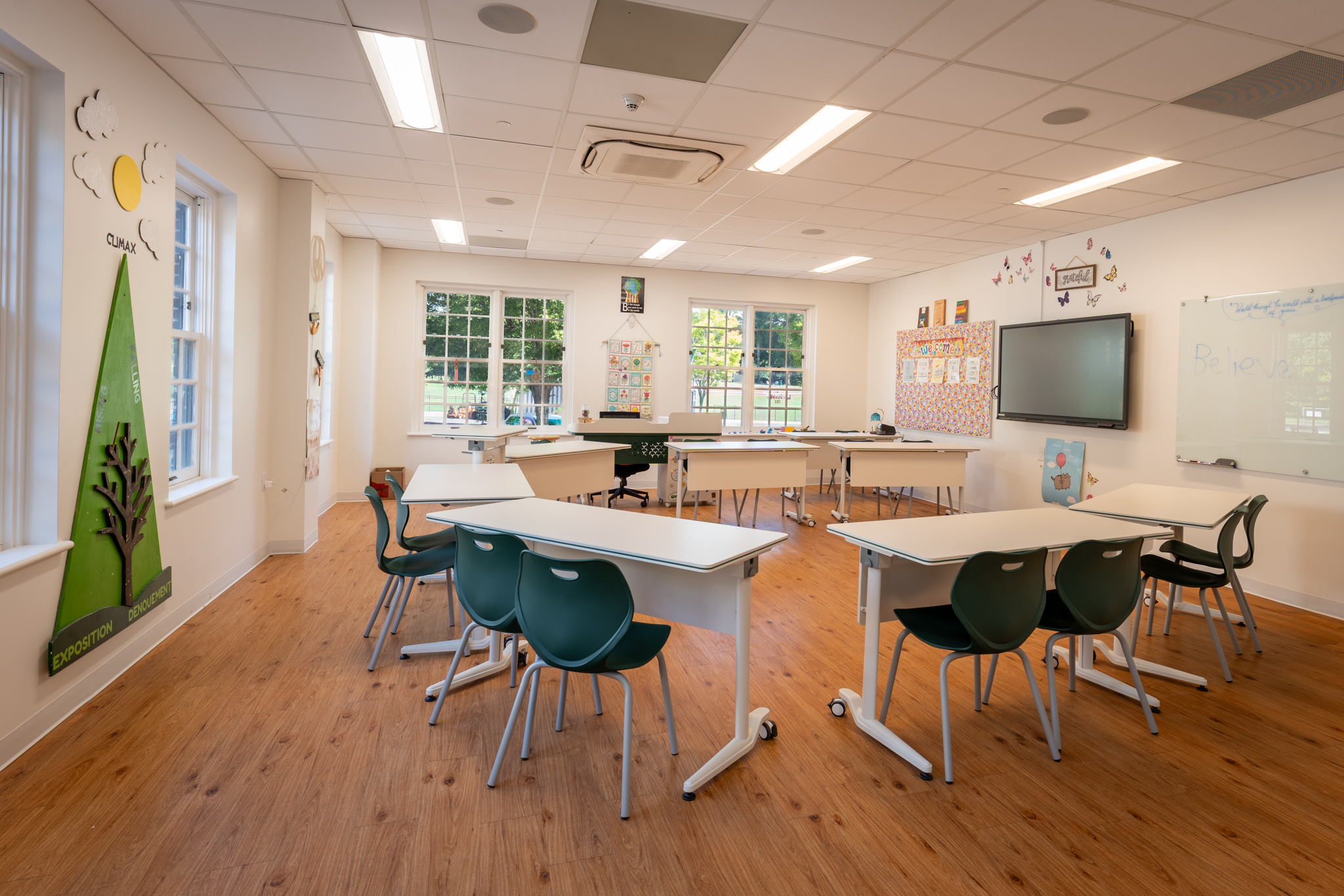 Flexible classroom with grouped white tables, green chairs, wood flooring, and a teaching screen