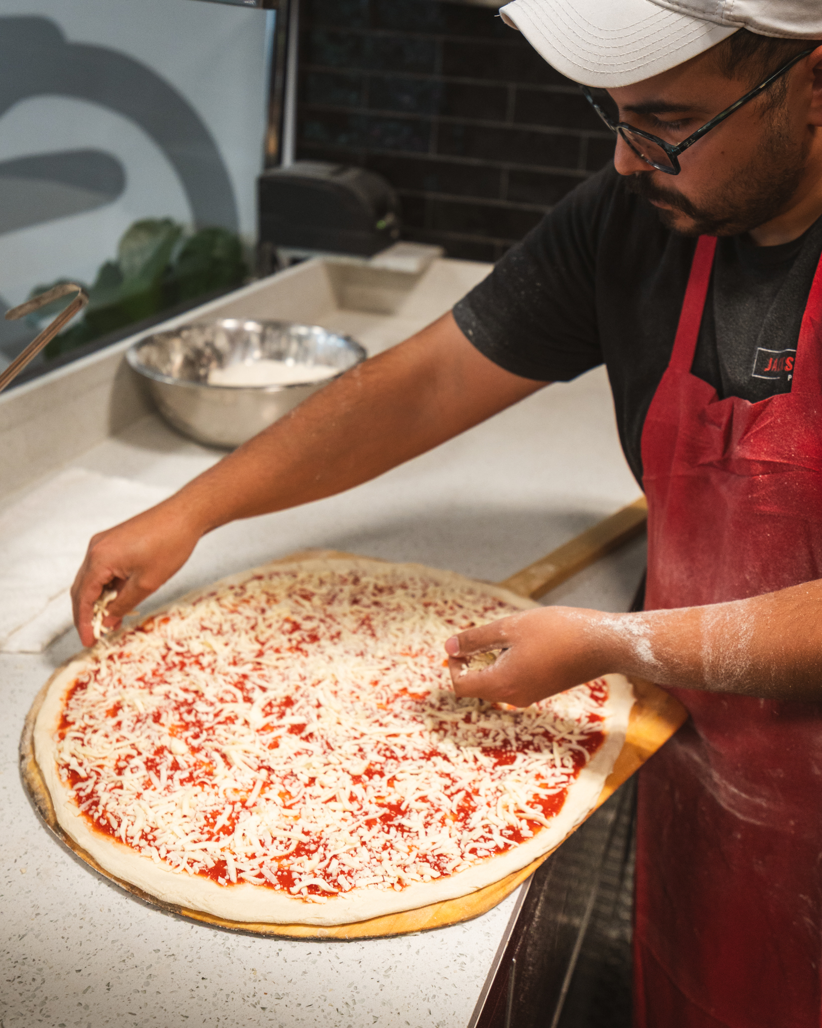 A pizza maker adding shredded cheese to a tomato-covered pizza before baking