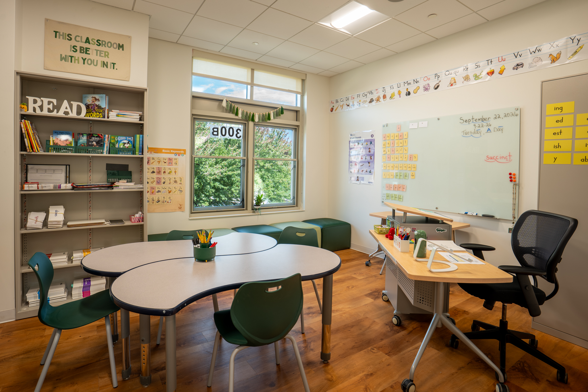 Grade-appropriate classroom with curved learning tables, green chairs, reading shelves, and a whiteboard