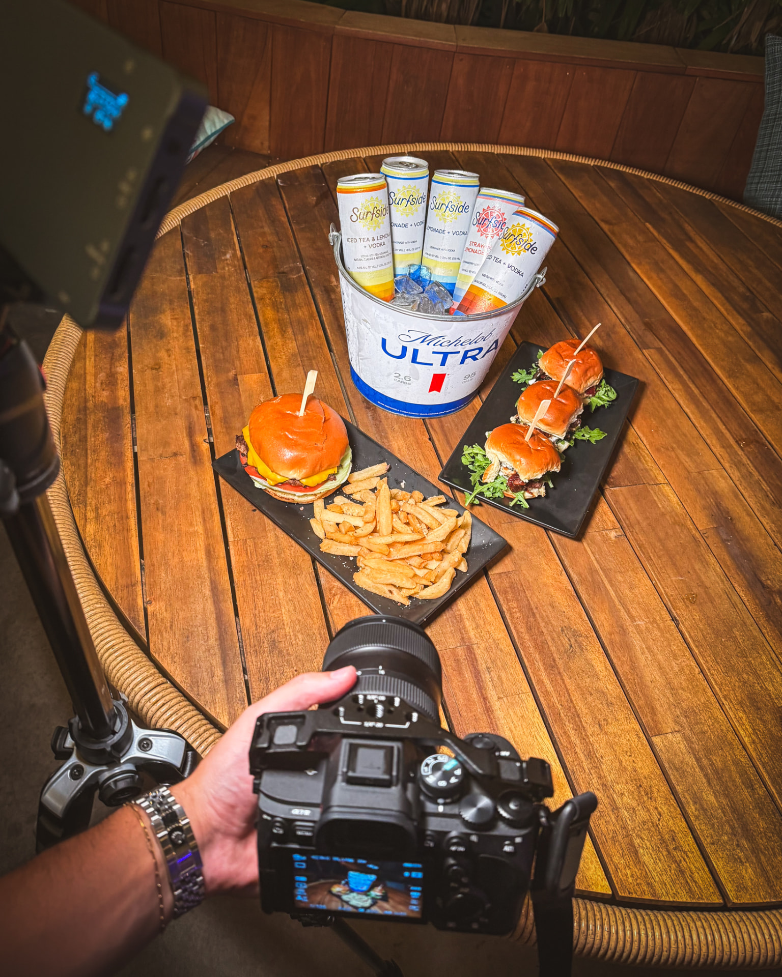 A photographer’s hand holding a camera over a table of burgers, fries, sliders, and drinks