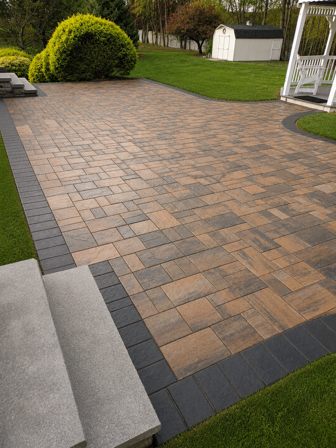 Warm-toned paver patio with a contrasting dark border beside a garden lawn