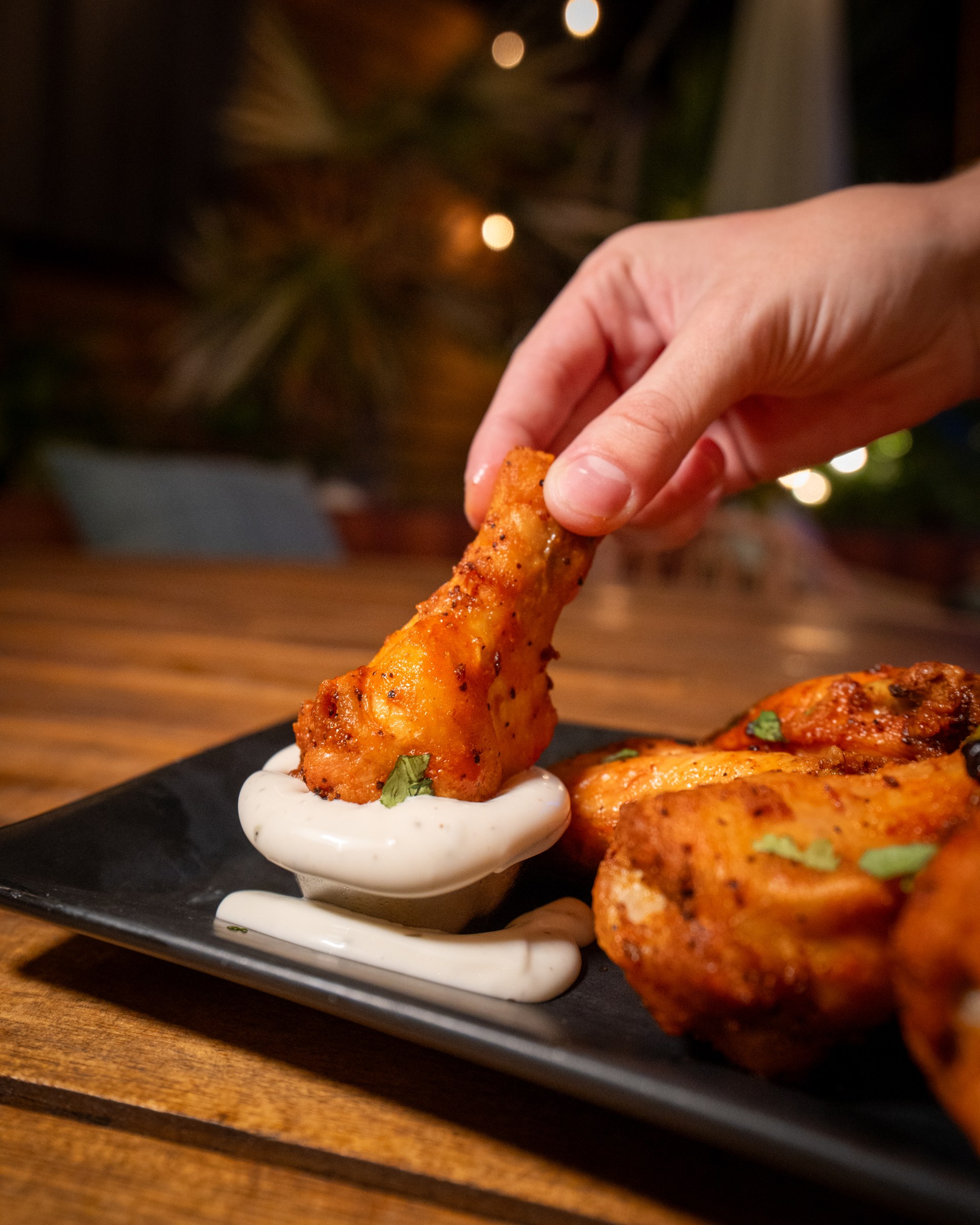 A chicken wing being dipped into sauce beside a plate of wings