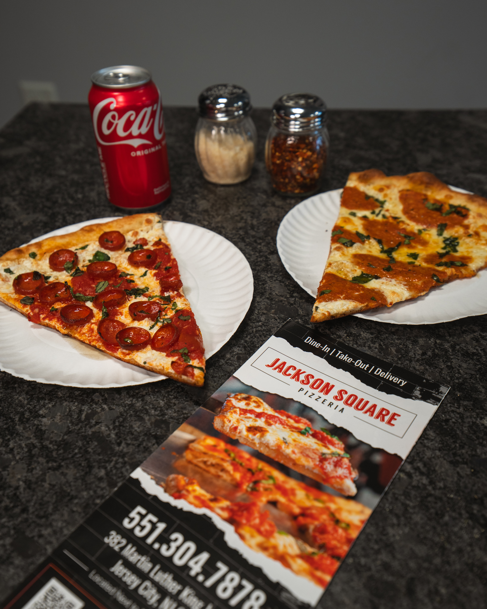 Pizza slices, a red soda can, and a Jackson Square Pizzeria menu arranged on the shop counter