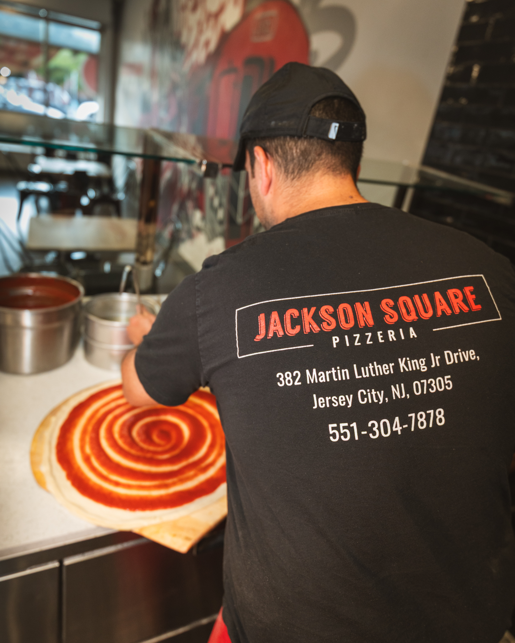 A pizza maker in a Jackson Square Pizzeria shirt spiraling tomato sauce over fresh dough