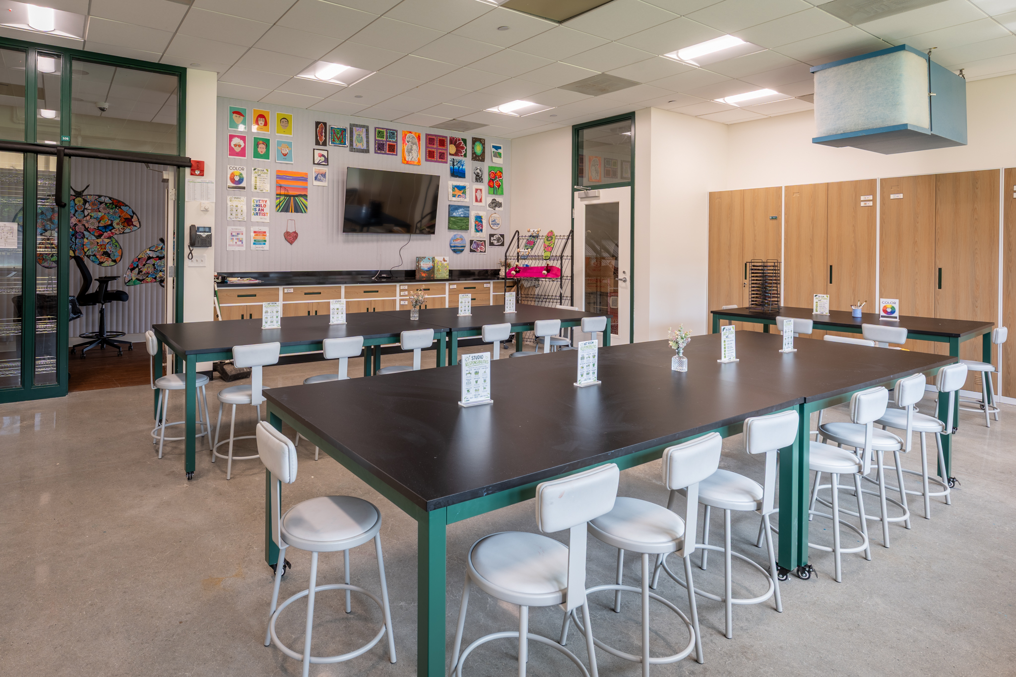 Completed school studio with a large black worktable, white stools, storage cabinetry, and green-framed glass doors